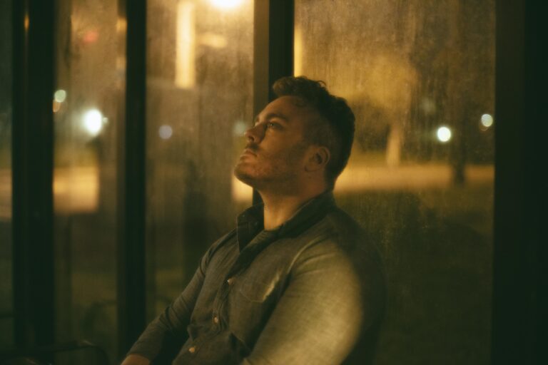 Homme assis la nuit dans un abribus, la tête appuyée contre la vitre embuée, le regard levé, éclairé par la lumière orangée des lampadaires