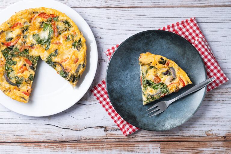 Frittata aux épinards, tomates et oignons rouges dans une assiette blanche, une part servie dans une assiette bleue avec une fourchette, sur une table en bois