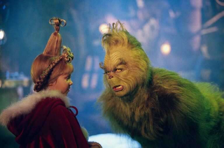 Le Grinch au pelage vert, l'air renfrogné, face à une petite fille aux cheveux relevés en chignon et en manteau rouge, dans le film de 2000