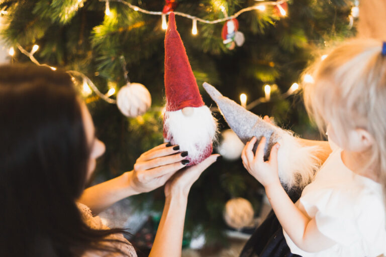 Une femme et une petite fille tenant deux figurines de lutins en feutrine à long bonnet, devant un sapin de Noël illuminé
