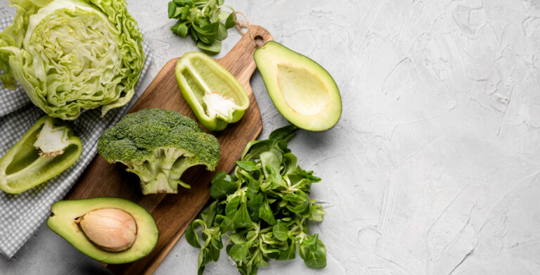 Légumes verts sur une planche en bois : demi-avocats, brocoli, poivron vert coupé, laitue et mâche, sur un plan de travail gris