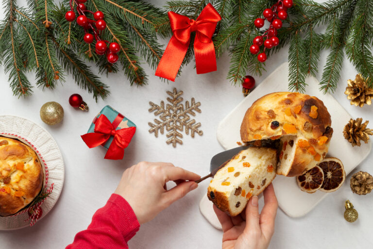 Panettone aux fruits confits coupé en parts sur une planche blanche, entouré de branches de sapin, de baies rouges, d'une pomme de pin et d'un petit cadeau