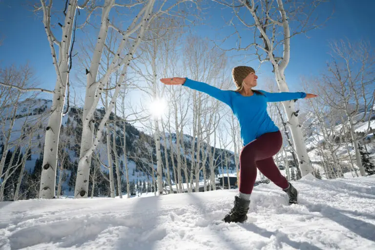Femme en tenue de sport faisant une posture de yoga dans la neige, entre des bouleaux, sous un grand soleil d'hiver en montagne