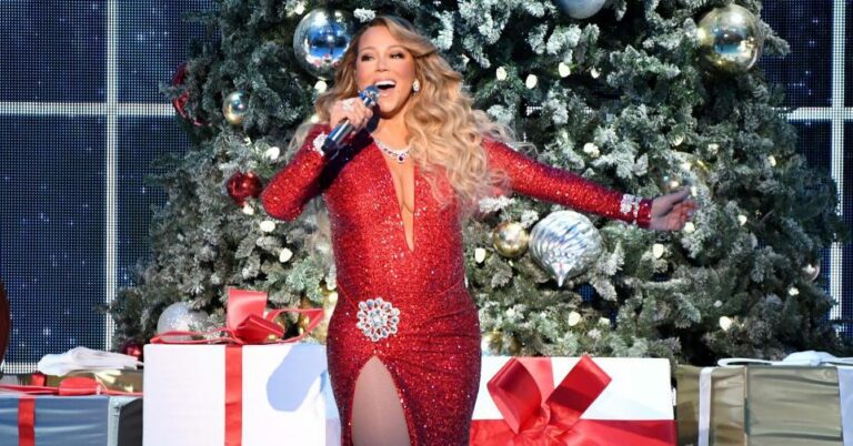 Mariah Carey en robe rouge à paillettes chantant au micro sur scène, devant un grand sapin de Noël décoré et des paquets cadeaux
