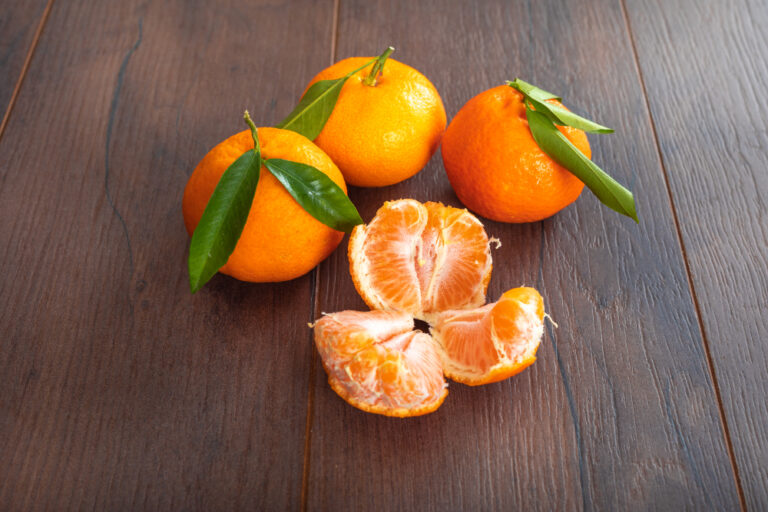 Trois petits agrumes orange avec leurs feuilles vertes sur une table en bois sombre, à côté d'un fruit épluché en quartiers