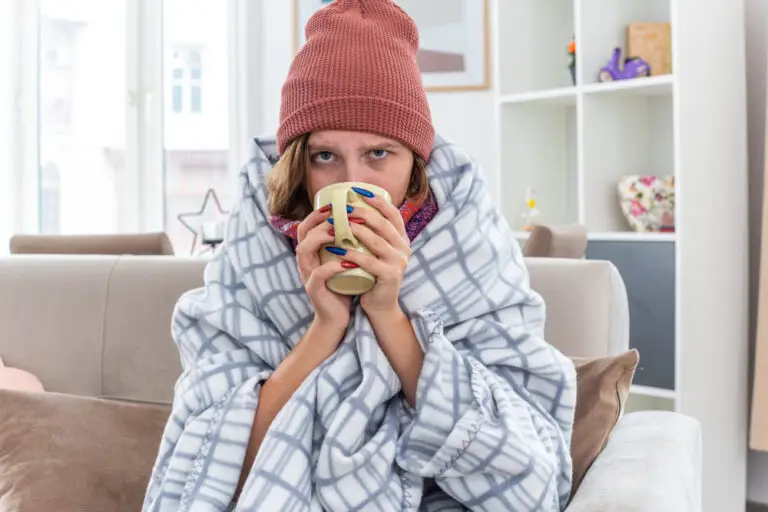 Jeune femme en bonnet de laine, enroulée dans un plaid à carreaux, buvant une boisson chaude dans son canapé, l'air enrhumé