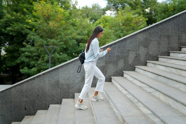 Jeune femme en jean blanc et chemise claire montant un grand escalier de pierre en extérieur, un café et un téléphone à la main