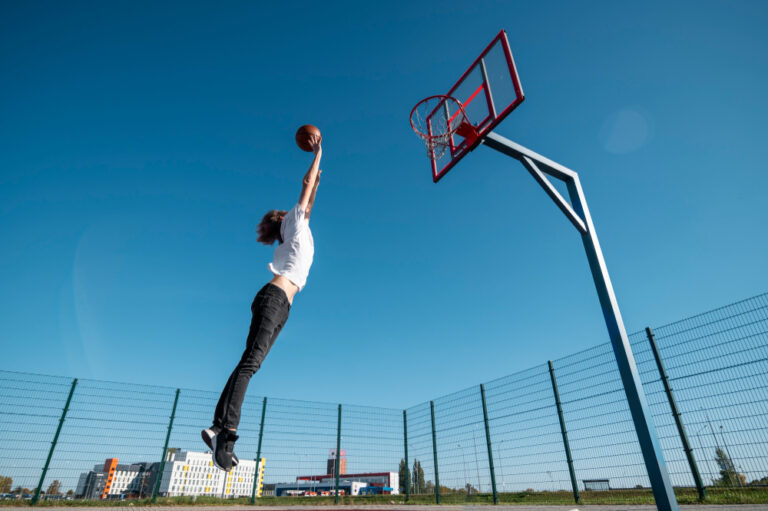 Jeune joueur sautant vers un panier de basket sur un terrain extérieur grillagé, sous un ciel bleu