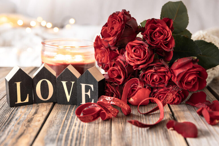 Lettres en bois formant le mot LOVE devant un bouquet de roses rouges, des rubans pailletés et une bougie allumée sur une table en bois
