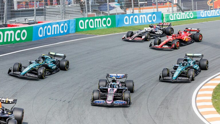 Six monoplaces de Formule 1 en peloton dans un virage du circuit de Zandvoort, bordé de panneaux publicitaires verts et bleus