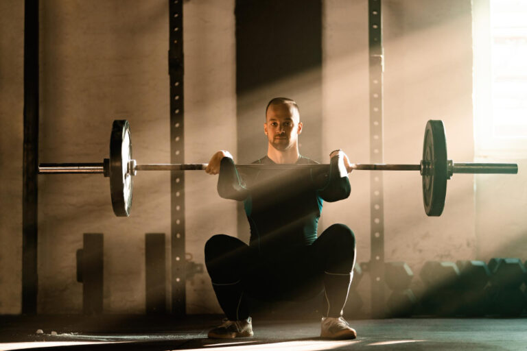 Homme faisant un squat avant avec une barre chargée dans une salle de sport, éclairé par un rayon de soleil