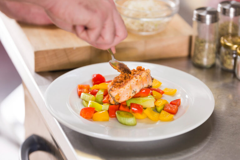 Un cuisinier dépose une chapelure sur un pavé de saumon servi sur des légumes colorés, poivrons et poireaux, dans une assiette blanche