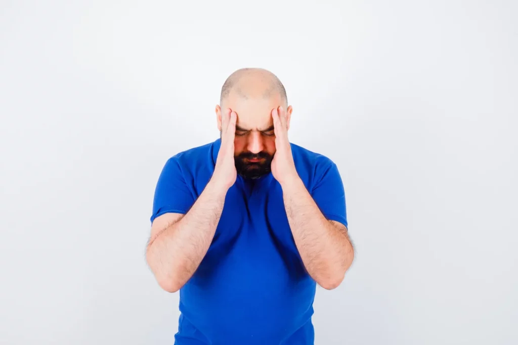 Homme barbu en t-shirt bleu, tête baissée, les deux mains plaquées sur les tempes, sur fond blanc