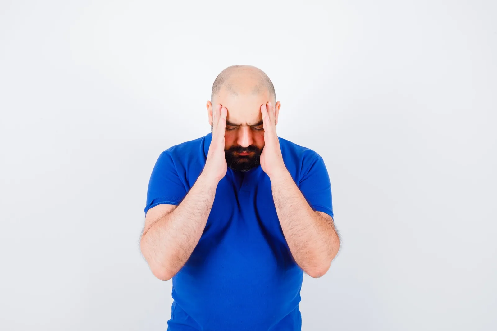 Homme barbu en t-shirt bleu, tête baissée, les deux mains plaquées sur les tempes, sur fond blanc