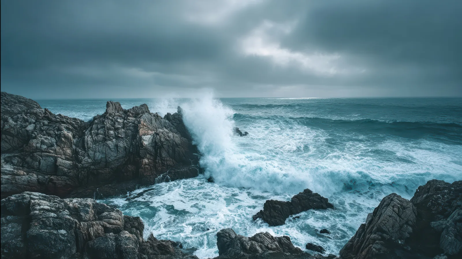 Vague qui se brise contre des rochers sous un ciel gris et chargé, mer agitée jusqu'à l'horizon
