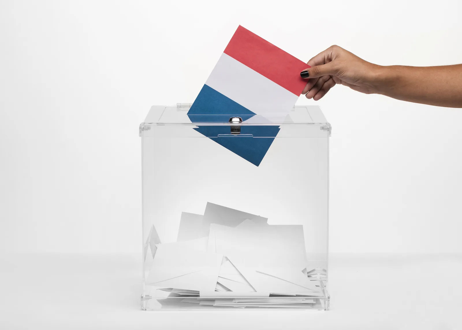 Main qui glisse un bulletin aux couleurs du drapeau français dans une urne transparente à moitié remplie d'enveloppes blanches
