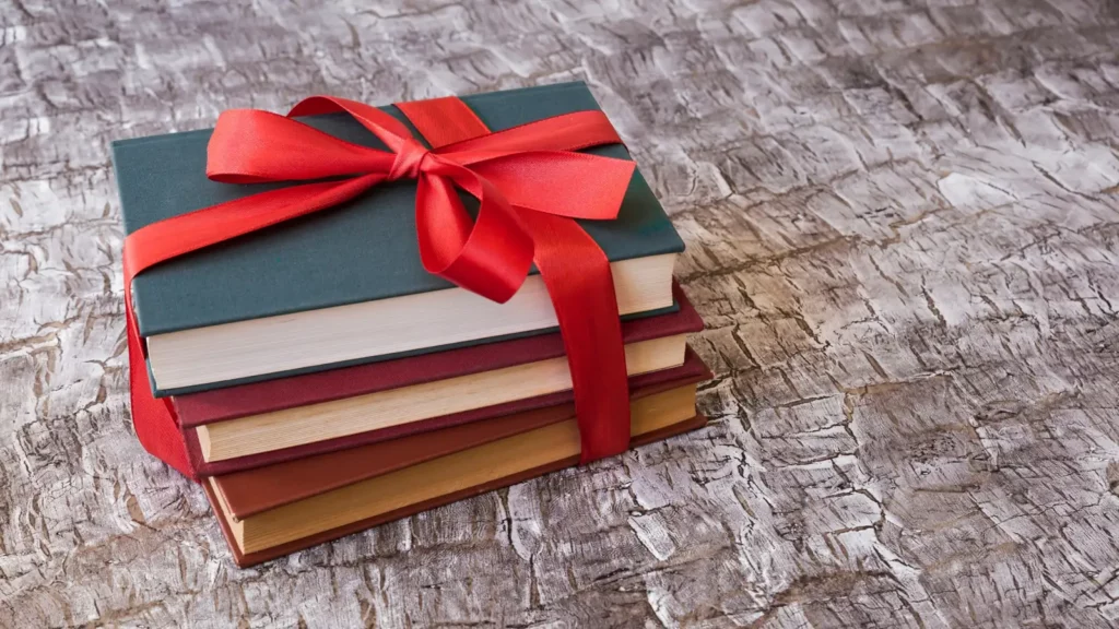 Pile de quatre livres reliés, vert, bordeaux et brun, nouée d'un large ruban rouge, posée sur une table en bois patiné