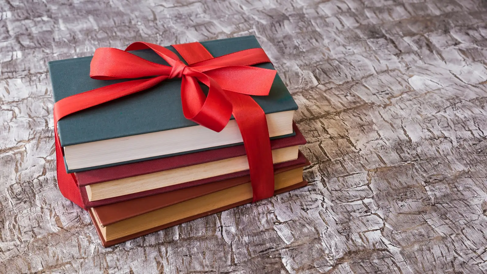 Pile de quatre livres reliés, vert, bordeaux et brun, nouée d'un large ruban rouge, posée sur une table en bois patiné